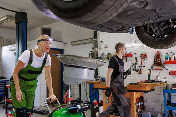 Woman mechanic using a vacuum-style oil extractor to remove engine oil from a car elevated on a hydraulic lift
