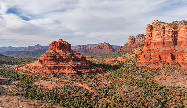 A sweeping aerial view of Bell Rock and Courthouse Butte near Sedona, Arizona, captures the vivid red sandstone formations that define this desert landscape. The contrasting green vegetation and layered red rocks under a softly clouded sky highlight the dramatic beauty of the American Southwest.